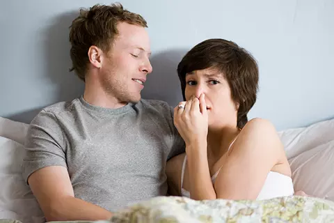 Ein Paar liegt im Bett und sie hält sich die Nase zu weil er Blähungen hat.