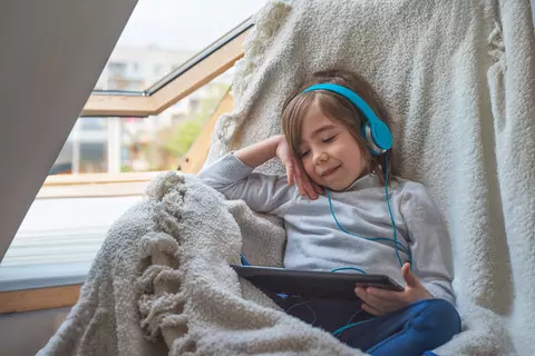 Kind sitzt mit Tablet und Kopfhörern mit einer Decke am offenen Fenster
