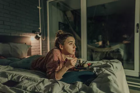 Frau liegt auf dem Bett, isst und schaut Fernsehen