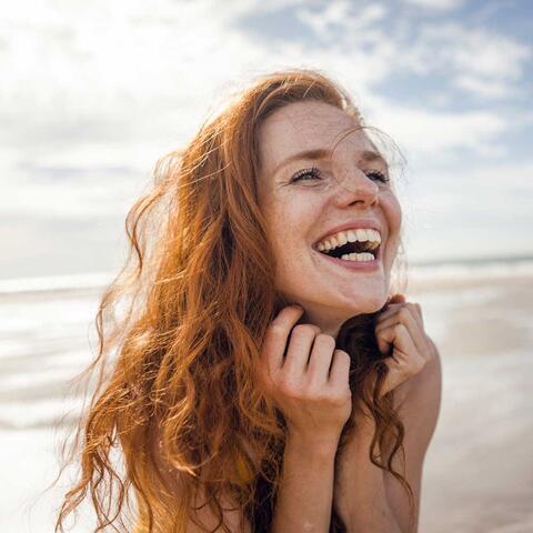 Junge Frau am Strand fasst sich in die Haare und lacht